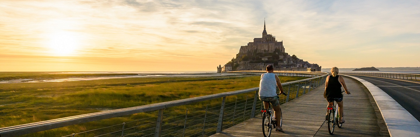 View Of The Mont Saint Michel Tidal Island In Normandy, France, At Sunset With A Couple Biking To The Town On The Wooden Jetty.